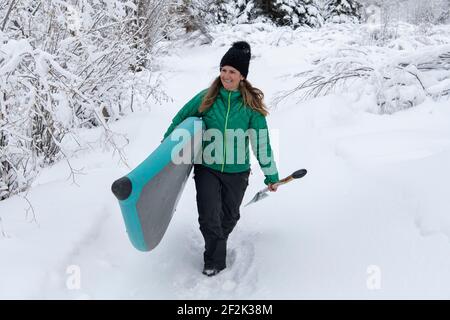 Lächelnde Frau in warmer Kleidung mit Paddelbrett beim Gehen Schneefeld Stockfoto