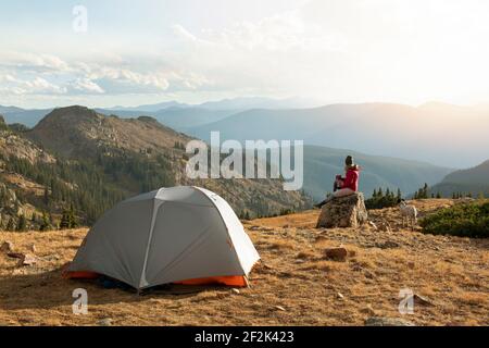 Frau auf Felsen sitzend, während sie mit Hund auf dem Berg zeltet Gegen den Himmel im Urlaub Stockfoto