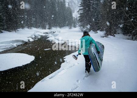 Rückansicht der Frau, die ein Paddelbrett trägt, während sie auf dem Schneefeld läuft Stockfoto