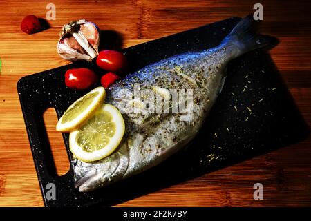 Roher dorado-Fisch oder Seebrasse mit Rosmarin, Limette, Knoblauch und Tomaten auf einem Schneidebrett. Stockfoto