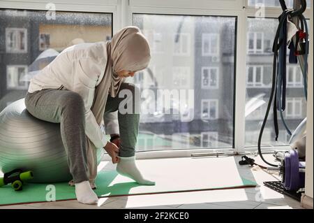 Die athletische arabische Muslimin in Hijab sitzt auf einem Fitnessball und zieht weiße Socken an, um sich für ein Workout vorzubereiten. Heimtraining. In Innenräumen. Stockfoto