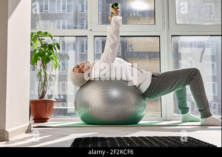 Charmante arabische muslimische Frau nett lächelnd an der Kamera, dabei Übungen an den Händen mit Hanteln, während auf einem Sportball liegen Stockfoto