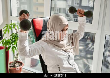 Porträt einer schönen sportlichen arabischen Frau im Kopftuch sitzend Auf einer Pressbank und beim Training mit eisernen Hanteln Zu Hause Stockfoto