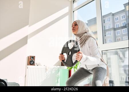 Arabische muslimische Frau mit Kopfschmuck trägt Hijab dabei gewichtete Kniebeugen mit einer Metallscheibe in den Händen - Home Workout-Konzept. Stockfoto