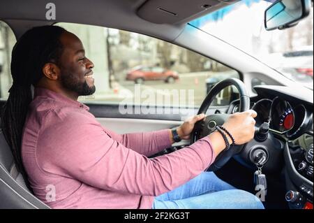 Ein glücklicher afroamerikanischer Mann mit den Dreadlocks fährt ein Auto, Seitenansicht von einem Beifahrersitz auf einen fröhlichen multirassischen Mann mit Lenkrad, kaufte ein Auto, lernte fahren Stockfoto