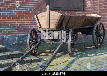 Ein brauner hölzerner Vintage-Wagen mit hölzernen Rädern, das Zugpendel nach vorne auf einem alten grauen Steinsteig mit Gras überwuchert gegen eine rote Ziegelwand w Stockfoto