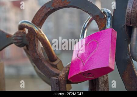 Ein rosafarbenes Vorhängeschloss mit geprägtem Herz, das an den schwarz gestrichenen rostigen halbrunden Elementen des Brückengeländers hängt. Konzept: Valentinstag, Unio Stockfoto