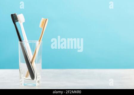 Ökologische Zahnbürste aus natürlichem Bambus und zwei Kunststoff-Zahnbürsten in Glas auf einem Holztisch, blauer Hintergrund. Nachhaltiger Lebensstil Stockfoto