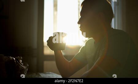 Silhouette eines jungen Mannes in den Sonnenstrahlen der Morgendämmerung trinkt heißen Kaffee, während zu Hause sitzen, Kamerabewegung Stockfoto