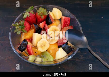 Natürlicher und gesunder Obstsalat mit Orange in einer transparenten Glasschale, auf einer rustikalen schwarzen Oberfläche. Gesunde Ernährung Konzept, Gesundes Leben. 45 Grad Stockfoto