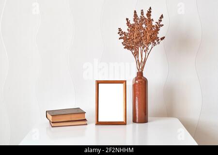 Inneneinrichtung. Hölzerne braune Rahmen Nachbau, Tonvase mit braunen getrockneten Blumen, Stapel von zwei alten kleinen Büchern auf einem weißen Couchtisch gegen ein w Stockfoto