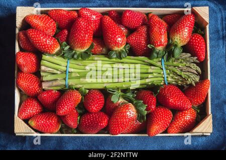 Blick von oben auf ein Holztablett mit einem Bündel von grünen wilden Spargel umgeben von frischen Erdbeeren auf einem blauen Stoff Textur Hintergrund. Stockfoto