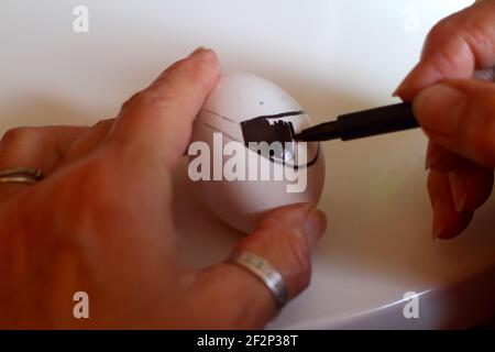 Die Hand einer Frau malt ein Osterei. Vorbereitung Ostern Dekorationen auf Multicolor Hintergrund. Sie zeichnet eine N95 Maske auf das Ei. Selektiver Fokus. Stockfoto