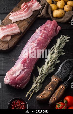 Roher Schweinelende mit Zutaten und Kräutern zum Backen, Salbei, Kartoffel-Set, auf schwarzem Holztischhintergrund Stockfoto