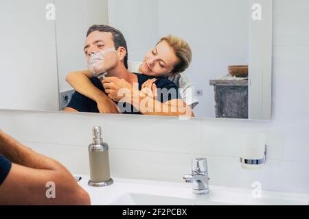Junges Paar zu Hause im Schlafzimmer, während Mann tun Und schneiden Sie Bart bereit, den Tag zu beginnen - Liebe Leute Indoor Freizeitbeschäftigung - Freundin umarmen Freund von hinten Und genießen Stockfoto