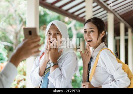 Zwei Mädchen in indonesischer High School Uniform mit schockierten Ausdrücken Wenn ein Junge einen Smartphone-Bildschirm zeigt Stockfoto