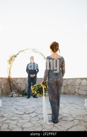 Eine Braut mit einem Blumenstrauß in der Hand geht an Der Bräutigam, der bei der Hochzeit auf sie wartet Arch Stockfoto