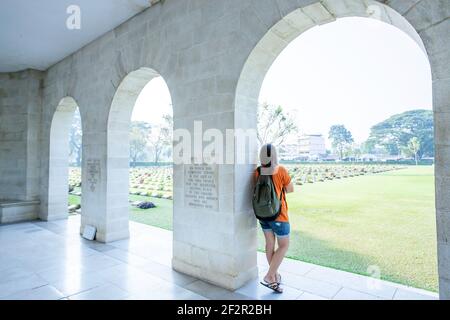 Rückansicht der jungen asiatischen Frau steht am gewölbten Eingang des Kanchanaburi Kriegsfriedhofs (Don Rak), während sie den Friedhof der Kriegsgräber betrachtet. Stockfoto