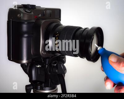 Nahaufnahme isolierte POV-Aufnahme einer Männerhand mit einem blauen Gummibläser, um Staub aus dem Glas eines Mittelformat-Filmkameraobjektivs auf einem Stativ zu entfernen. Stockfoto