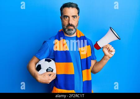 Mann mittleren Alters mit Bart und grauen Haaren Fußball Hooligan hält Ball und mit Megaphon im Schockgesicht, suchen skeptisch und sarkastisch, überrascht w Stockfoto