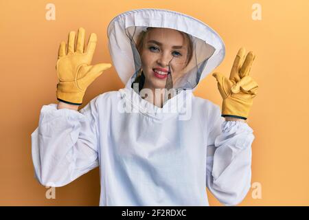 Schöne blonde kaukasische Frau trägt schützende Imker Uniform zeigen und Zeigt mit den Fingern Nummer acht nach oben und lächelt zuversichtlich und Ha Stockfoto