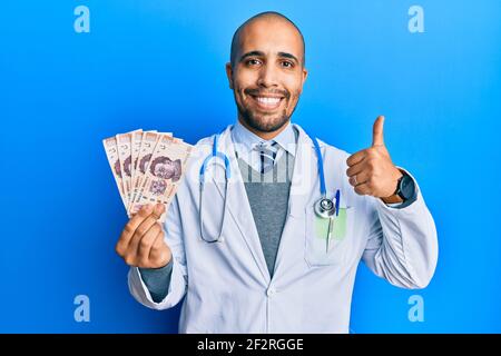 Hispanische Erwachsene Arzt Mann trägt medizinische Uniform hält 500 mexikanische Pesos lächelnd glücklich und positiv, Daumen nach oben tun ausgezeichnet und Zustimmung Zeichen Stockfoto