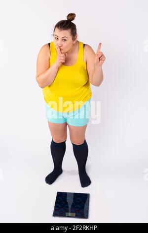 Eine kaukasische prall gekleidete Frau steht vor einer elektronischen Waage und zeigt ein Schild, das ihren Zeigefinger leise vor ihr Gesicht hält. Stockfoto