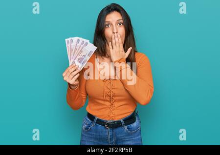 Schöne hispanische Frau hält russische 500 Rubel Banknoten über den Mund mit der Hand, schockiert und Angst vor Fehler. Überrascht Ausdruck Stockfoto