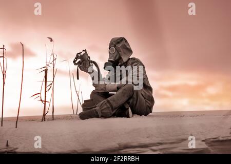 Ein Mann in Gasmaske und Schutzanzug sitzt auf dem Sand und hält die Gasmaske eines toten Freundes in der Hand. Stockfoto