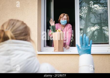 Soziale Distanzierung durch Coronavirus covid-19 Pandemiesperre. Ältere Frau mit Gesichtsmaske winkte aus dem Fenster zu ihrer erwachsenen Tochter, sie lieferte Essen Stockfoto