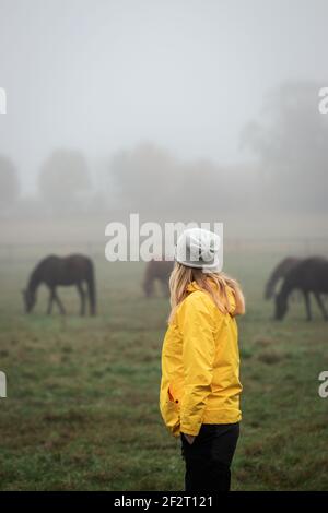 Frau genießen Blick auf Pferd auf der Weide im Herbst nebligen Morgen. Tourist trägt gelben Regenmantel und Hut. Ruhige Szene mit Tieren Stockfoto