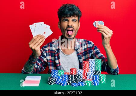 Junge hispanic Mann spielen Poker mit Karten und Casino-Chips ragen Zunge aus glücklich mit lustigen Ausdruck. Stockfoto