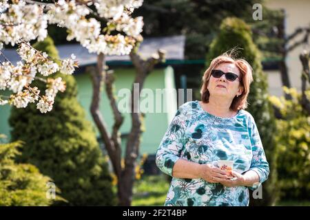 Glückliche ältere Frau genießt sonnigen Tag unter blühenden Kirschbaum. Ältere Erwachsene Frau trinkt Kaffee und entspannt im Garten im Frühling Stockfoto