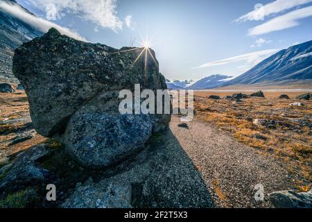 Sunstar am Rande eines großen Felsbrockens, der auf dem Boden eines abgelegenen arktischen Tals sitzt. Akshayuk Pass, Baffin Island, Kanada. Sonniger Tag des arktischen Sommers in Stockfoto