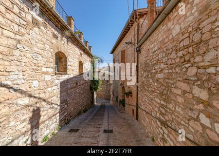Eine schmale gepflasterte Straße schrägt sich zwischen warmen Steinhäusern und hohen Mauerwänden in der historischen Hügelstadt Spello, Umbrien, Italien. Stockfoto