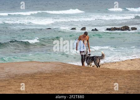 Mann kommt aus dem Meer in der Nähe von Famara Strand auf Lanzarote mit Surfbrett unter dem Arm. Sein Hund wartet auf ihn am Strand Stockfoto