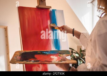Frau malt in ihrem Atelier. Rote und blaue Acrylfarbe auf Leinwand. Palette mit Ölfarben in Künstlerhand Stockfoto