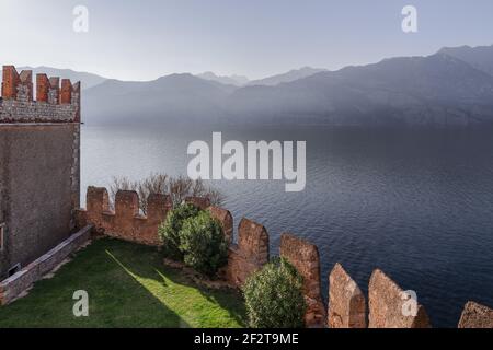 Zackige Mauern des Scaliger-Schlosses von Malcesine überblicken den Gardasee, mit nebligen Bergen im Hintergrund, Venetien, Italien. Stockfoto