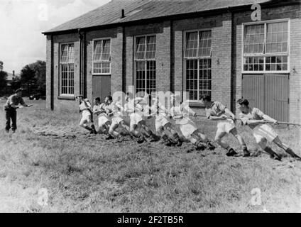 1950s, historischer, nationaler Dienst nach dem Krieg, ein Armeeoffizier, der acht junge Soldaten in ihren Turnschuhen und Armeestiefeln Anweisungen gibt, die bei einer Tauziehen-Übung auf Gras vor ihren Kasernen, England, Großbritannien, an einem Seil ziehen. Stockfoto