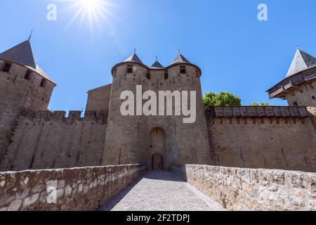 Eine Steinbrücke führt zu den Zwillings-Torwehren des Chateau Comtal unter einem Sonnenstrahl in der Cité von Carcassonne, Occitanie, Frankreich. Stockfoto