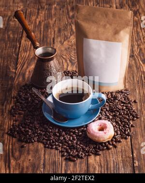 Blaue Tasse Kaffee, Donuts, Bohnen, türkische Kaffeekanne und Bastelpapierbeutel auf Holzhintergrund Stockfoto