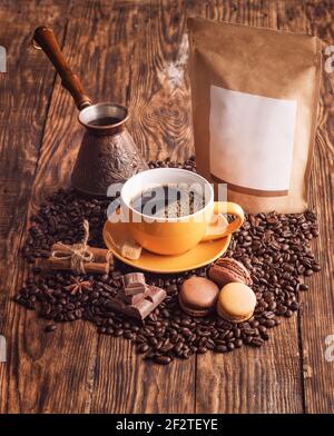 Gelbe Tasse Kaffee, Makronen, Bohnen, türkische Kaffeekanne und Bastelpapierbeutel auf Holzbackgroun Stockfoto