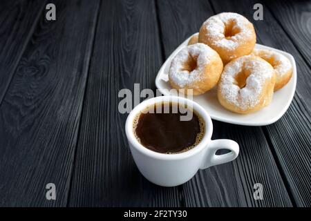 Schwarzer Kaffee in der weißen Tasse und hausgemachter Donut mit Puderzucker auf dem schwarzen Holzhintergrund. Speicherplatz kopieren. Nahaufnahme. Stockfoto