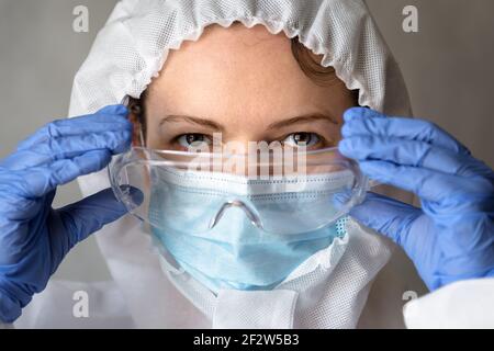 Ärztin in PPE-Anzug Nahaufnahme, Krankenschwester setzt auf medizinische Brille während COVID-19 Pandemie, Porträt einer Ärztin trägt Schutzmaske wegen Stockfoto