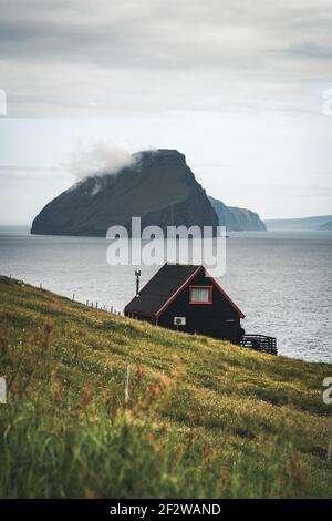 Schwarzes Haus auf dem berühmten faroese Witches Finger Trail und der Insel Koltur im Hintergrund. Sandavagur Dorf, Vagar Insel, Färöer Inseln, Dänemark Stockfoto