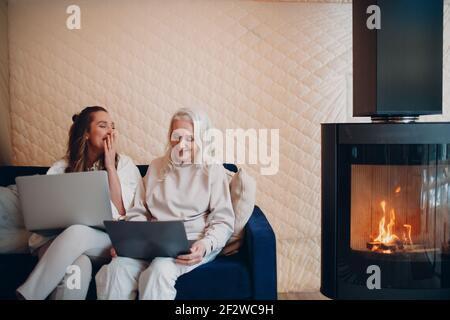 Ältere und junge Frau mit Computer-Laptop auf dem Sofa. Mama und Tochter sitzen am Kamin drinnen in Datscha. Stockfoto