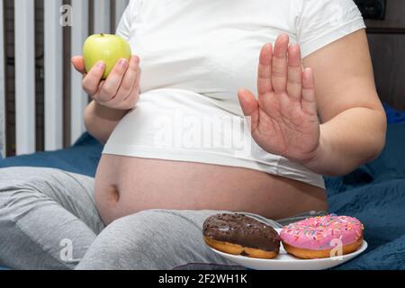 Nahaufnahme einer schwangeren Frau, die einen grünen Apfel in den Händen hält und Donuts ablehnt. Gesunde Ernährung während der Schwangerschaft Konzept. Gewichtskontrolle. Stockfoto