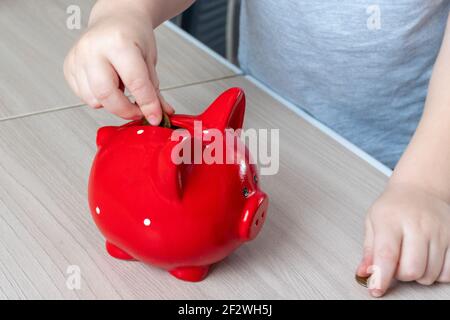 Kinderhand legt eine Münze in ein rotes Sparschwein auf einem hölzernen Hintergrund, Draufsicht, Kopierraum. Sparkonzept Stockfoto