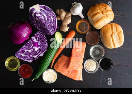 Asiatisches Lachsbrötchen mit Sesamsalat Zutaten: Rohe Lachsfilets, Gemüse, Hamburger-Brötchen und Gewürze auf dunklem Holz Stockfoto
