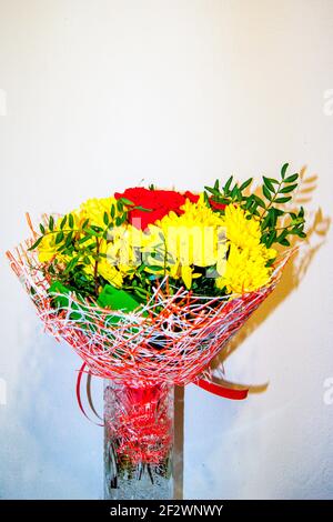 Ein Bouquet aus gelben Chrysanthemen und roten Rosen in der Verpackung steht in einer Vase gegen die weiße Wand. Geschenk. Eine Liebeserklärung. Blumen Stockfoto